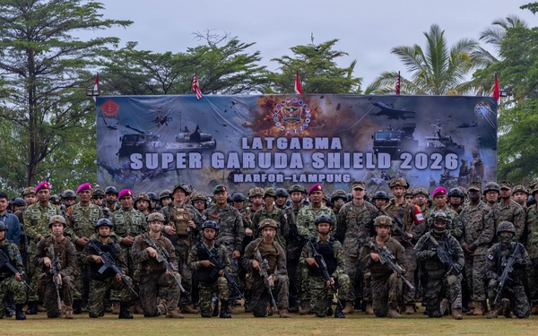 A large group of military personnel from multiple nations pose for a formal group photograph at the 'Super Garuda Shield 2026' joint military exercise held at Marinir Lampung. The participants are arranged in rows on a grass field, wearing various military uniforms and combat gear. A large banner identifying the exercise is prominently displayed behind the assembled troops.