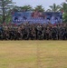 A large group of military personnel from multiple nations pose for a formal group photograph at the 'Super Garuda Shield 2026' joint military exercise held at Marinir Lampung. The participants are arranged in rows on a grass field, wearing various military uniforms and combat gear. A large banner identifying the exercise is prominently displayed behind the assembled troops.