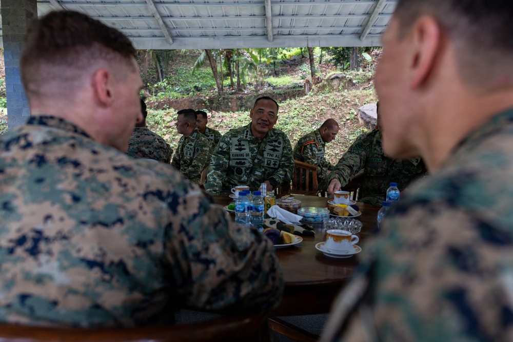 Military personnel from at least two different armed forces are gathered around a table in an outdoor covered area, appearing to be in a meeting or social engagement. The central figure wears a uniform with 'TNI AL' and 'KANANG BR' name tape, indicating Indonesian Navy (Tentara Nasional Indonesia Angkatan Laut) affiliation. Two foreground figures wear MARPAT (Marine Pattern) camouflage consistent with U.S. Marines.