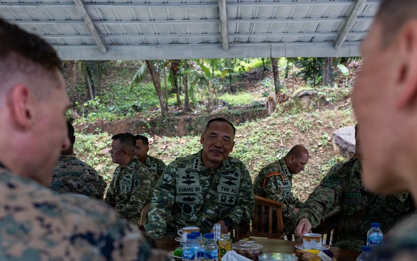 Military personnel from at least two different armed forces are gathered around a table in an outdoor covered area, appearing to be in a meeting or social engagement. The central figure wears a uniform with 'TNI AL' and 'KANANG BR' name tape, indicating Indonesian Navy (Tentara Nasional Indonesia Angkatan Laut) affiliation. Two foreground figures wear MARPAT (Marine Pattern) camouflage consistent with U.S. Marines.