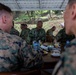 Military personnel from at least two different armed forces are gathered around a table in an outdoor covered area, appearing to be in a meeting or social engagement. The central figure wears a uniform with 'TNI AL' and 'KANANG BR' name tape, indicating Indonesian Navy (Tentara Nasional Indonesia Angkatan Laut) affiliation. Two foreground figures wear MARPAT (Marine Pattern) camouflage consistent with U.S. Marines.