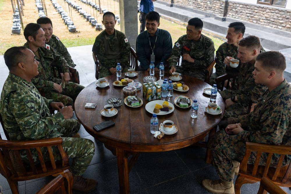 A multinational military gathering shows personnel from multiple nations seated around a large round wooden table in what appears to be an outdoor covered pavilion. Soldiers wearing uniforms with visible Japanese, South Korean, and US Marine Corps insignia are engaged in conversation alongside what appears to be Indonesian or other Southeast Asian military personnel. The setting suggests a formal diplomatic or joint exercise meeting with refreshments served.