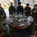 A multinational military gathering shows personnel from multiple nations seated around a large round wooden table in what appears to be an outdoor covered pavilion. Soldiers wearing uniforms with visible Japanese, South Korean, and US Marine Corps insignia are engaged in conversation alongside what appears to be Indonesian or other Southeast Asian military personnel. The setting suggests a formal diplomatic or joint exercise meeting with refreshments served.