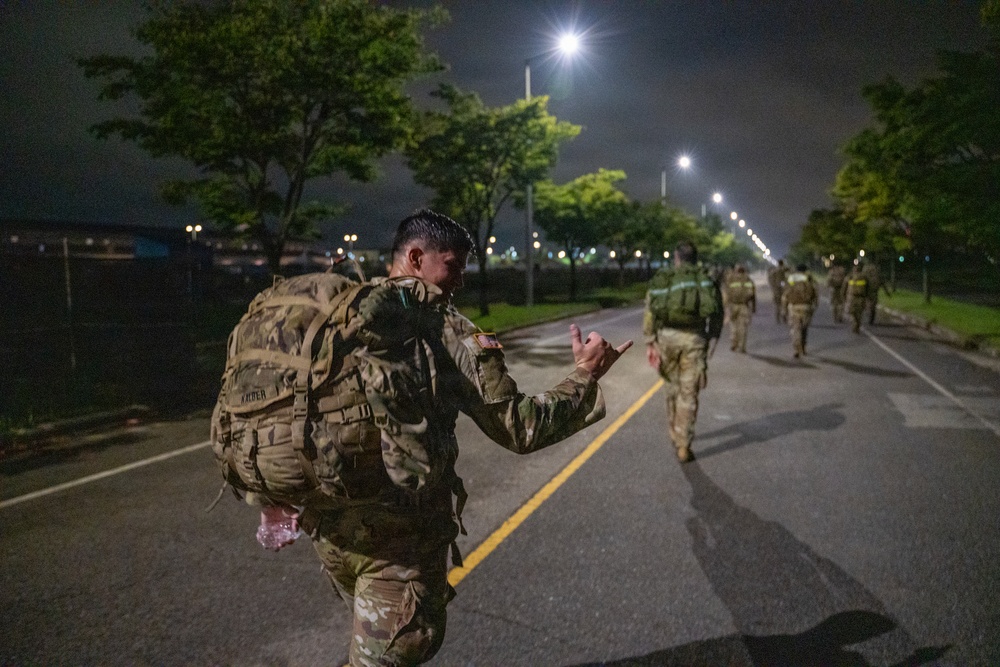 A group of military personnel in camouflage uniforms are conducting a ruck march along a paved road at night. The foreground soldier, whose name tape appears to read 'KAISER' or similar, is carrying a large tactical backpack and making a hand gesture toward fellow soldiers ahead. The scene is illuminated by street lights lining the road.
