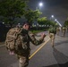 A group of military personnel in camouflage uniforms are conducting a ruck march along a paved road at night. The foreground soldier, whose name tape appears to read 'KAISER' or similar, is carrying a large tactical backpack and making a hand gesture toward fellow soldiers ahead. The scene is illuminated by street lights lining the road.