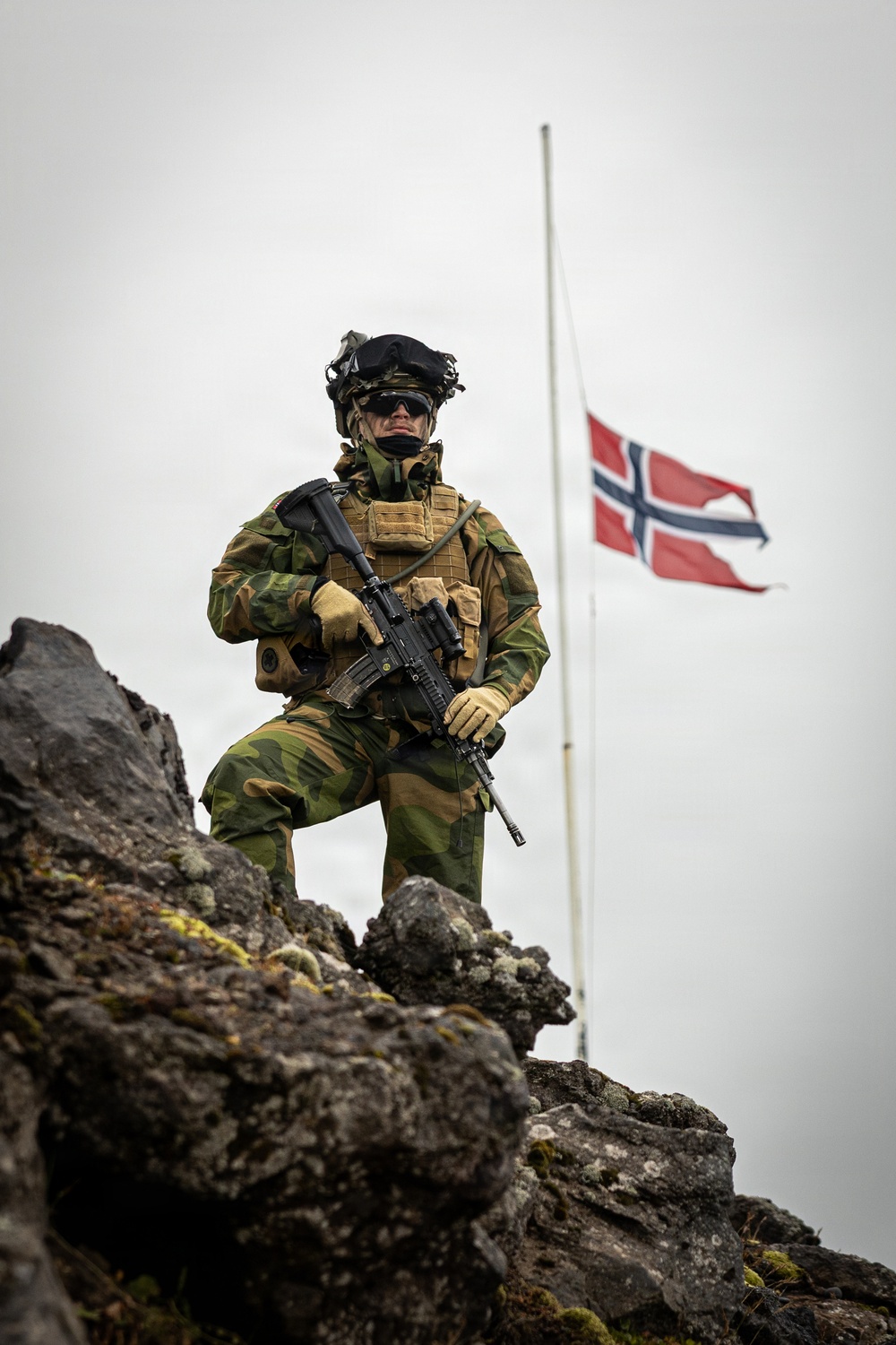 A fully equipped soldier in Norwegian military camouflage stands on rocky terrain holding an assault rifle, with the Norwegian flag flying on a flagpole in the background. The low-angle shot emphasizes the soldier's imposing stance against an overcast sky. The image appears to be a professional military photograph, likely taken in Norway or a Nordic environment given the rocky, lichen-covered terrain.