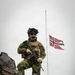 A fully equipped soldier in Norwegian military camouflage stands on rocky terrain holding an assault rifle, with the Norwegian flag flying on a flagpole in the background. The low-angle shot emphasizes the soldier's imposing stance against an overcast sky. The image appears to be a professional military photograph, likely taken in Norway or a Nordic environment given the rocky, lichen-covered terrain.