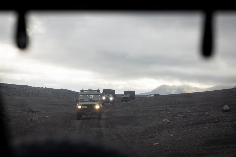 The image is taken from inside a vehicle looking out through what appears to be a rear window or opening, showing a convoy of military-style vehicles traversing a dark, barren volcanic or highland terrain. The lead vehicle has its headlights on and is driving along a dirt track toward the camera. Mountains obscured by clouds are visible in the background under an overcast sky.