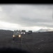 The image is taken from inside a vehicle looking out through what appears to be a rear window or opening, showing a convoy of military-style vehicles traversing a dark, barren volcanic or highland terrain. The lead vehicle has its headlights on and is driving along a dirt track toward the camera. Mountains obscured by clouds are visible in the background under an overcast sky.