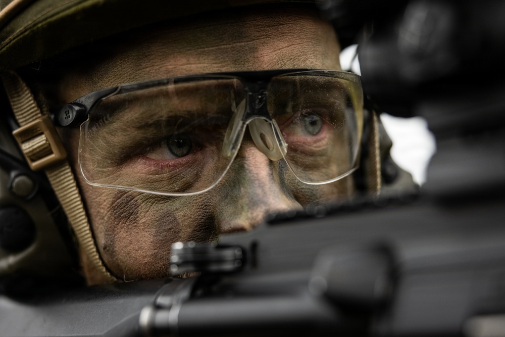 A close-up photograph of a male soldier or tactical operator with camouflage face paint applied to his skin. He is wearing ballistic/protective clear-lens eyewear, a tactical helmet, and communication ear protection. He appears to be aiming or sighting down a firearm, with intense focus visible in his eyes.