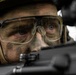 A close-up photograph of a male soldier or tactical operator with camouflage face paint applied to his skin. He is wearing ballistic/protective clear-lens eyewear, a tactical helmet, and communication ear protection. He appears to be aiming or sighting down a firearm, with intense focus visible in his eyes.