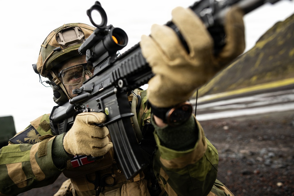 A Norwegian military soldier is shown in a tactical aiming position with an assault rifle equipped with an optical red dot sight. The soldier is wearing full combat gear including a camouflage helmet, protective eyewear, tactical gloves, and a Norwegian flag patch on the left arm. The image appears to be taken during a military exercise or training operation in a rocky, outdoor environment.
