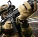 A Norwegian military soldier is shown in a tactical aiming position with an assault rifle equipped with an optical red dot sight. The soldier is wearing full combat gear including a camouflage helmet, protective eyewear, tactical gloves, and a Norwegian flag patch on the left arm. The image appears to be taken during a military exercise or training operation in a rocky, outdoor environment.