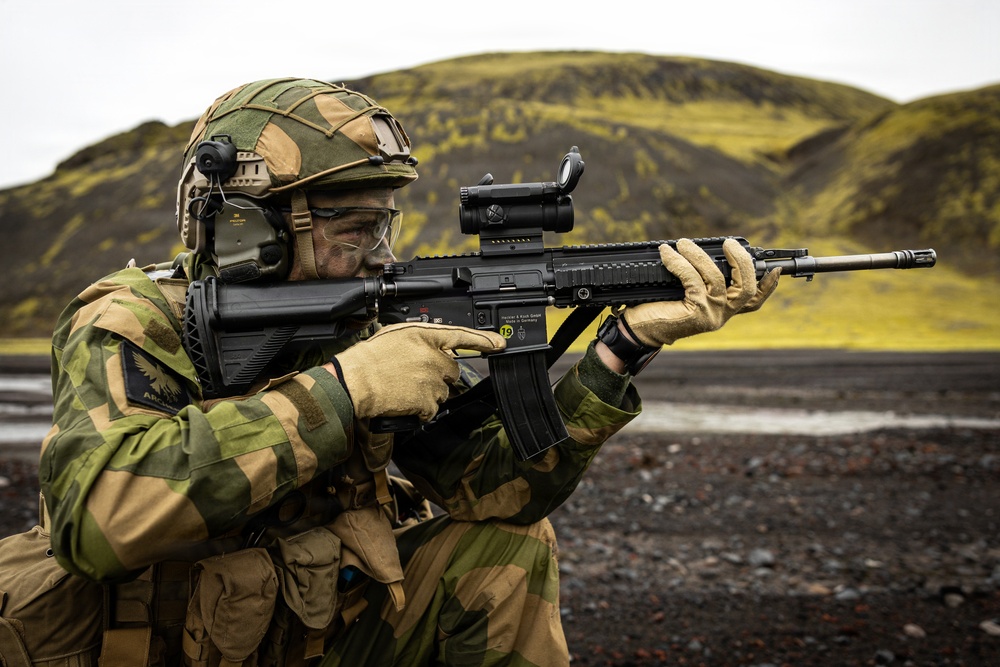 A soldier in camouflage uniform is aiming a rifle in an outdoor environment with volcanic black terrain and moss-covered hills in the background. The soldier is equipped with full tactical gear including a helmet, hearing protection, ballistic goggles, and tactical gloves. The weapon appears to be an HK33 or similar Heckler &amp; Koch rifle with an optic sight mounted on top.
