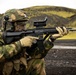 A soldier in camouflage uniform is aiming a rifle in an outdoor environment with volcanic black terrain and moss-covered hills in the background. The soldier is equipped with full tactical gear including a helmet, hearing protection, ballistic goggles, and tactical gloves. The weapon appears to be an HK33 or similar Heckler &amp; Koch rifle with an optic sight mounted on top.