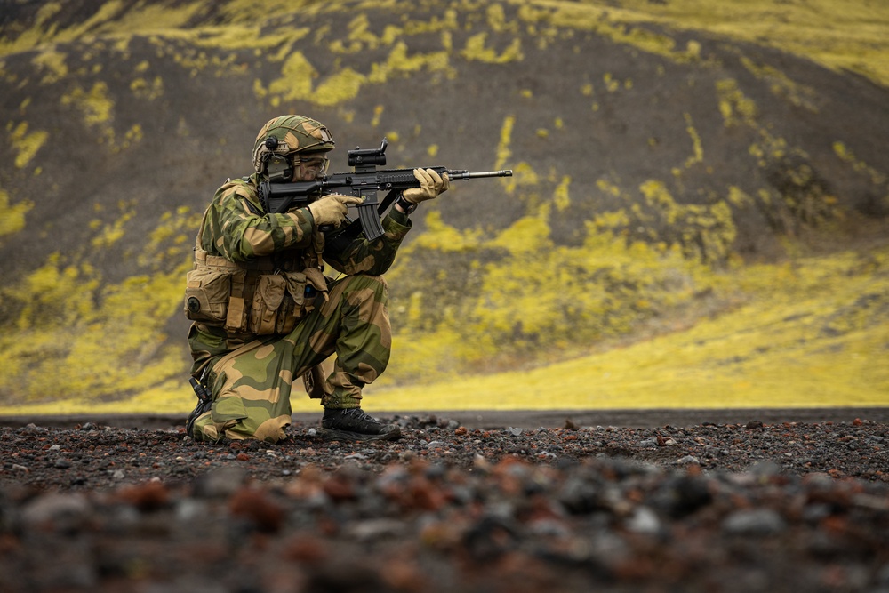 A soldier in full combat gear is kneeling in a low shooting stance on dark volcanic terrain, aiming a rifle toward the right of the frame. The background features dramatic yellow-green moss-covered volcanic hills, consistent with Icelandic or Nordic highland landscapes. The image appears to be a professional military training or promotional photograph.
