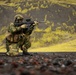 A soldier in full combat gear is kneeling in a low shooting stance on dark volcanic terrain, aiming a rifle toward the right of the frame. The background features dramatic yellow-green moss-covered volcanic hills, consistent with Icelandic or Nordic highland landscapes. The image appears to be a professional military training or promotional photograph.