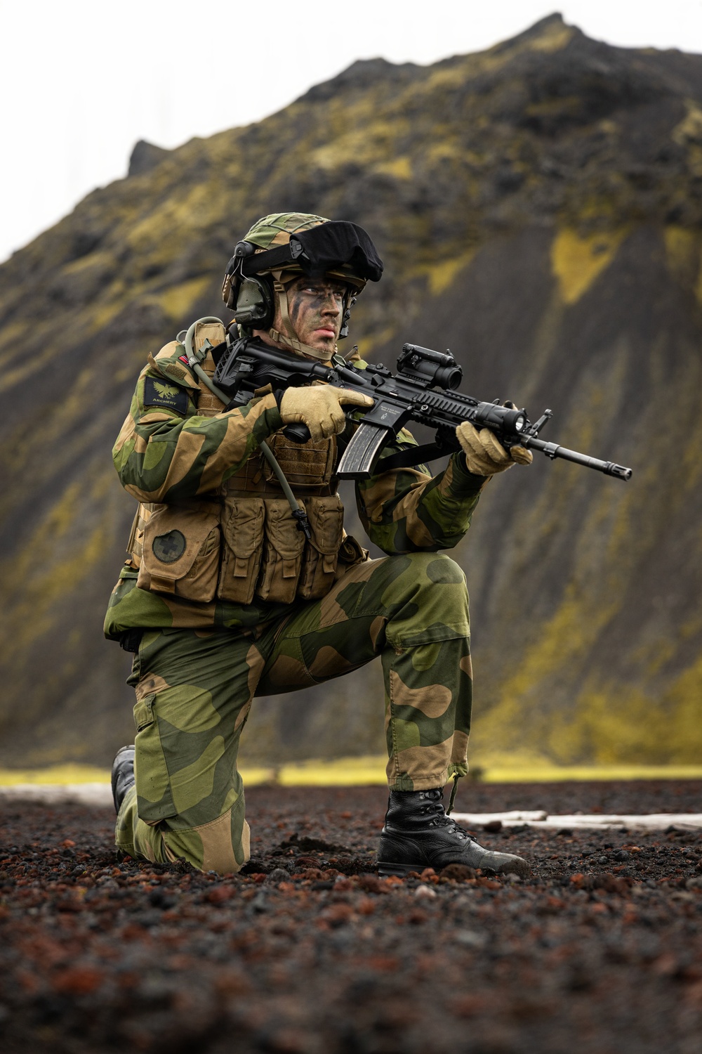 A soldier in full combat gear is kneeling in a low-ready firing position on dark volcanic terrain. The individual is equipped with camouflage uniform, tactical vest, helmet, and is aiming a rifle with an optical sight. The dramatic mountainous background with moss-covered slopes suggests a Nordic or Icelandic environment.
