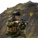 A soldier in full combat gear is kneeling in a low-ready firing position on dark volcanic terrain. The individual is equipped with camouflage uniform, tactical vest, helmet, and is aiming a rifle with an optical sight. The dramatic mountainous background with moss-covered slopes suggests a Nordic or Icelandic environment.