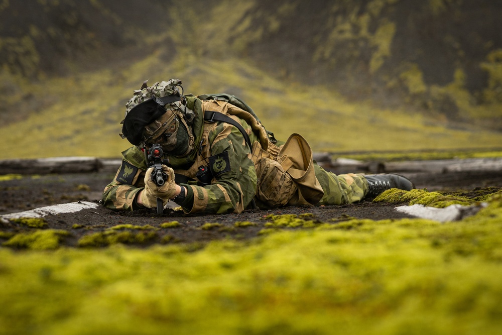 A soldier in camouflage uniform is lying prone on dark volcanic ground, aiming a rifle with a red dot sight. The setting appears to be a Nordic or sub-Arctic environment, characterized by moss-covered lava fields and green mountainous terrain in the background. The soldier is fully equipped with tactical gear including a backpack, helmet with attachments, face covering, and gloves.