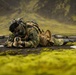 A soldier in camouflage uniform is lying prone on dark volcanic ground, aiming a rifle with a red dot sight. The setting appears to be a Nordic or sub-Arctic environment, characterized by moss-covered lava fields and green mountainous terrain in the background. The soldier is fully equipped with tactical gear including a backpack, helmet with attachments, face covering, and gloves.