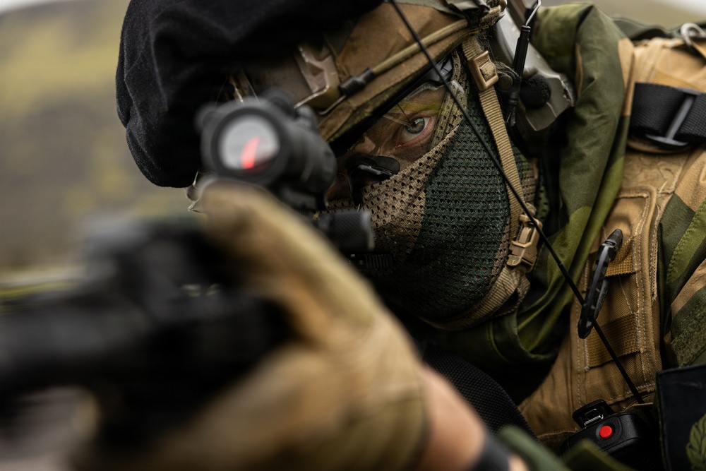 A heavily equipped soldier or tactical operator is shown in close-up, aiming a firearm fitted with a red dot optic sight directly toward the camera. The individual is wearing camouflage gear, a tactical helmet, a mesh face covering, and has green face paint applied. The image is a high-contrast, shallow depth-of-field photograph emphasizing the operator's focused eye and weapon sight.