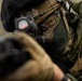 A heavily equipped soldier or tactical operator is shown in close-up, aiming a firearm fitted with a red dot optic sight directly toward the camera. The individual is wearing camouflage gear, a tactical helmet, a mesh face covering, and has green face paint applied. The image is a high-contrast, shallow depth-of-field photograph emphasizing the operator's focused eye and weapon sight.