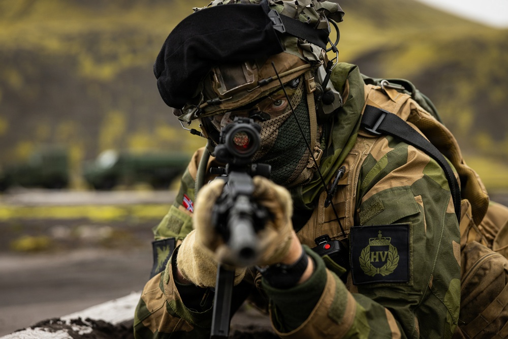 A soldier in full combat gear is aiming a rifle directly at the camera in a tactical position. The soldier wears a black beret, camouflage uniform, face covering, and tactical vest with identifiable unit patches. The background shows a rugged outdoor terrain with green hillsides.