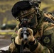 A soldier in full combat gear is aiming a rifle directly at the camera in a tactical position. The soldier wears a black beret, camouflage uniform, face covering, and tactical vest with identifiable unit patches. The background shows a rugged outdoor terrain with green hillsides.