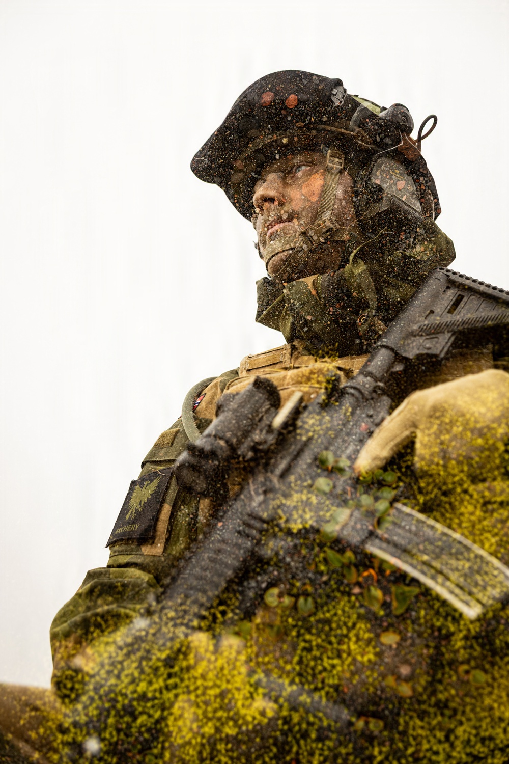A heavily equipped soldier is photographed from a low angle, covered in mud, debris, and vegetation, suggesting intense field conditions. The soldier is wearing full tactical gear including a combat helmet with mounted equipment, body armor, and carrying an assault rifle. A unit patch is visible on the left arm, and the overall scene conveys active military training or combat operations in harsh environmental conditions.