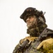 A heavily equipped soldier is photographed from a low angle, covered in mud, debris, and vegetation, suggesting intense field conditions. The soldier is wearing full tactical gear including a combat helmet with mounted equipment, body armor, and carrying an assault rifle. A unit patch is visible on the left arm, and the overall scene conveys active military training or combat operations in harsh environmental conditions.