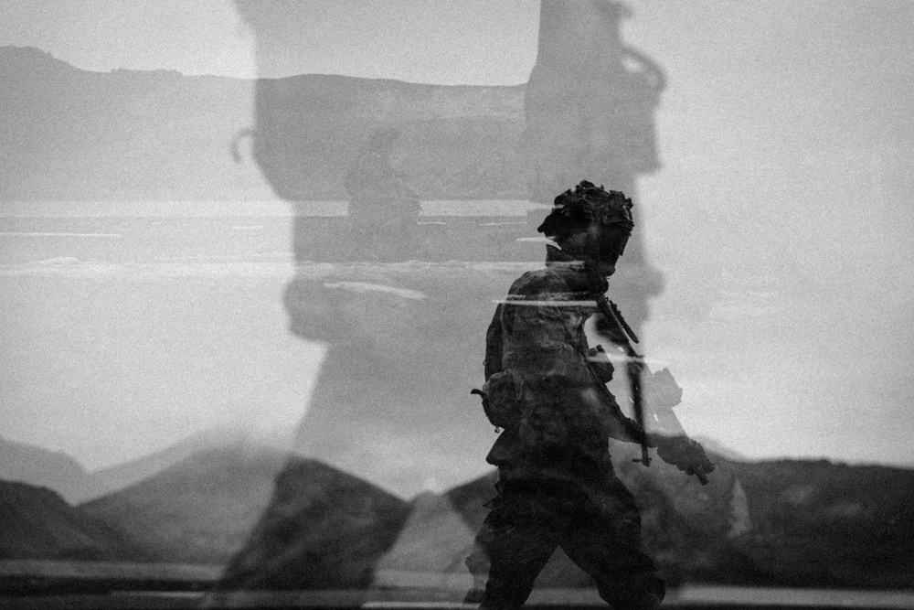 A black-and-white double-exposure or composite photograph showing a soldier in full combat gear walking against a backdrop that includes what appears to be a large military vehicle or equipment silhouette. The layered exposure creates a ghostly, surreal aesthetic with overlapping imagery of a desert or arid landscape. The image appears to be an artistic or editorial military photograph emphasizing scale and atmosphere.