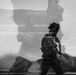 A black-and-white double-exposure or composite photograph showing a soldier in full combat gear walking against a backdrop that includes what appears to be a large military vehicle or equipment silhouette. The layered exposure creates a ghostly, surreal aesthetic with overlapping imagery of a desert or arid landscape. The image appears to be an artistic or editorial military photograph emphasizing scale and atmosphere.