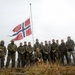 A group of approximately 14 military personnel in camouflage uniforms pose for a group photo on elevated terrain beneath a Norwegian flag flying from a tall flagpole. The overcast sky and rugged coastal landscape in the background suggest a cold, northern environment. The personnel appear to be from a joint or multinational military unit, wearing cold-weather gear including beanie hats and gloves.