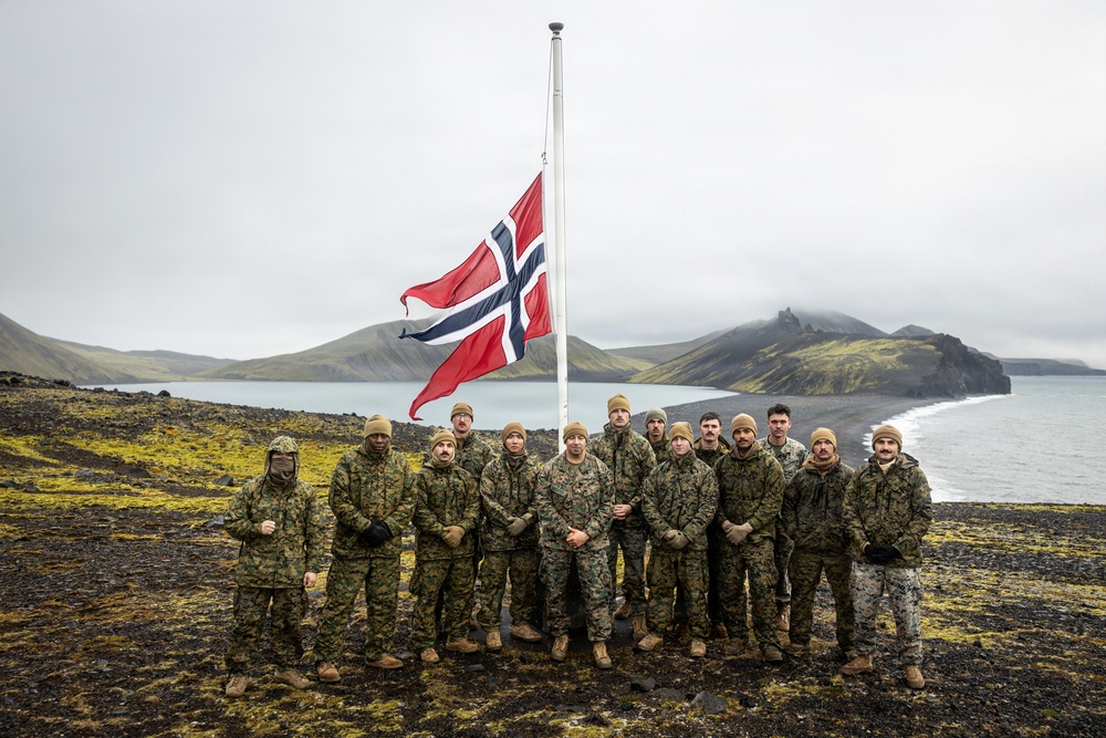 A group of approximately 13 military personnel in camouflage uniforms pose for a group photo in front of a Norwegian flag on a flagpole. The setting appears to be a remote, rugged coastal landscape with black volcanic terrain, green moss, and a body of water with mountains in the background, consistent with Iceland or a Nordic location. The overcast sky and cold-weather gear suggest harsh environmental conditions.