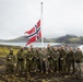 A group of approximately 13 military personnel in camouflage uniforms pose for a group photo in front of a Norwegian flag on a flagpole. The setting appears to be a remote, rugged coastal landscape with black volcanic terrain, green moss, and a body of water with mountains in the background, consistent with Iceland or a Nordic location. The overcast sky and cold-weather gear suggest harsh environmental conditions.