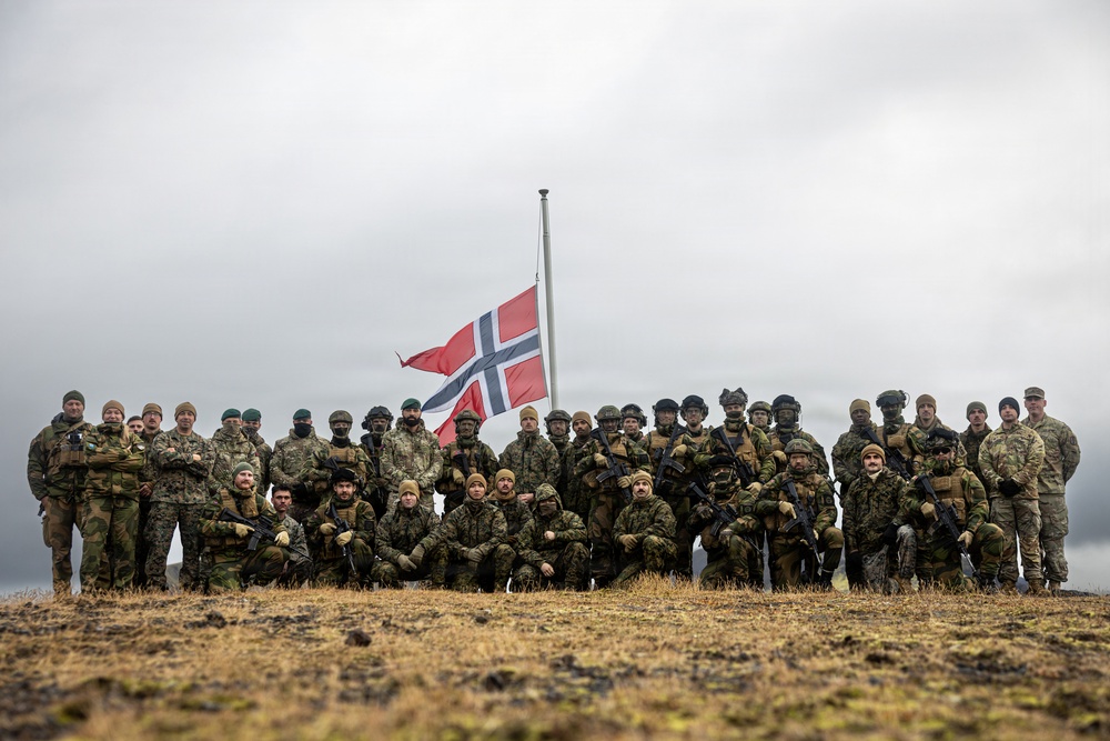 A large group of military personnel, approximately 30-40 individuals, pose for a group photograph on an elevated, open terrain under an overcast sky. A Norwegian flag is prominently displayed on a flagpole in the center background. The personnel are wearing various camouflage uniforms and tactical gear, suggesting a multinational military exercise or operation.