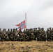 A large group of military personnel, approximately 30-40 individuals, pose for a group photograph on an elevated, open terrain under an overcast sky. A Norwegian flag is prominently displayed on a flagpole in the center background. The personnel are wearing various camouflage uniforms and tactical gear, suggesting a multinational military exercise or operation.