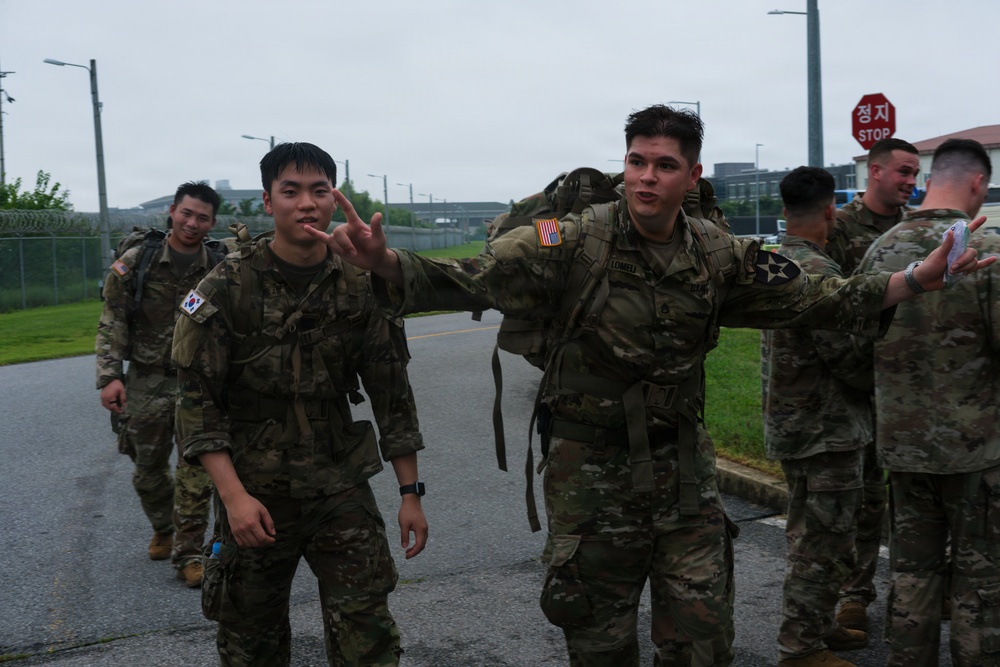 A group of soldiers in camouflage uniforms are walking along a road on what appears to be a military installation in South Korea. Two soldiers in the foreground — one wearing a South Korean flag patch and one wearing a U.S. flag patch with a 2nd Infantry Division insignia — are posing and gesturing toward the camera. The overcast weather and wet road suggest recent or ongoing rain.