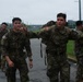 A group of soldiers in camouflage uniforms are walking along a road on what appears to be a military installation in South Korea. Two soldiers in the foreground — one wearing a South Korean flag patch and one wearing a U.S. flag patch with a 2nd Infantry Division insignia — are posing and gesturing toward the camera. The overcast weather and wet road suggest recent or ongoing rain.