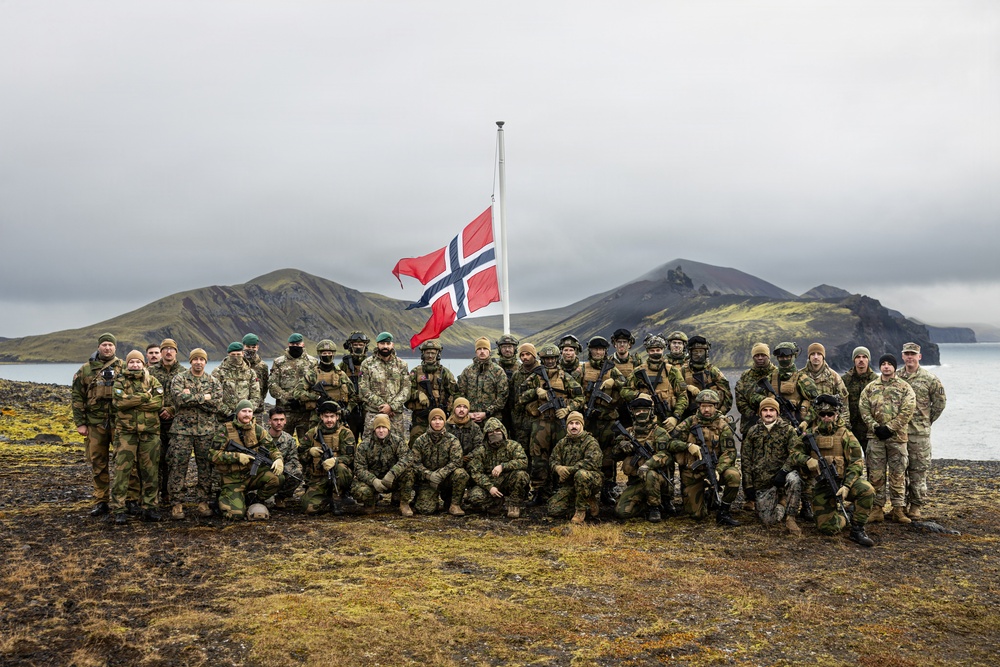 A large group of military personnel in various camouflage uniforms pose for a group photograph in front of a Norwegian flag on a flagpole. The setting appears to be a remote, rugged coastal landscape with volcanic hills and a body of water visible in the background. The group includes both standing and kneeling rows of soldiers, many carrying rifles.