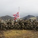 A large group of military personnel in various camouflage uniforms pose for a group photograph in front of a Norwegian flag on a flagpole. The setting appears to be a remote, rugged coastal landscape with volcanic hills and a body of water visible in the background. The group includes both standing and kneeling rows of soldiers, many carrying rifles.