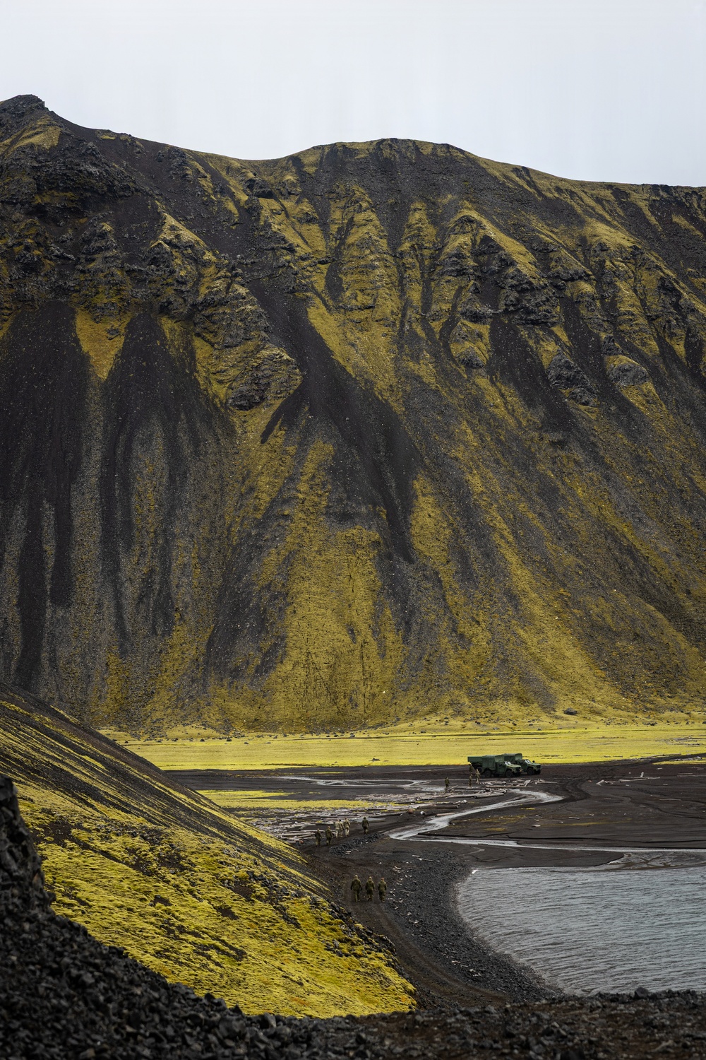 A dramatic volcanic landscape featuring a large dark mountain covered in vivid yellow-green moss or lichen, with black erosion channels running down its slopes. In the foreground, a black sand or volcanic ash valley contains a winding stream, a small group of people on foot, and a green military-style vehicle parked near the base of the mountain. The overcast sky and stark color contrast between the dark volcanic rock and bright vegetation create a striking, otherworldly atmosphere.