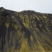 A dramatic volcanic landscape featuring a large dark mountain covered in vivid yellow-green moss or lichen, with black erosion channels running down its slopes. In the foreground, a black sand or volcanic ash valley contains a winding stream, a small group of people on foot, and a green military-style vehicle parked near the base of the mountain. The overcast sky and stark color contrast between the dark volcanic rock and bright vegetation create a striking, otherworldly atmosphere.