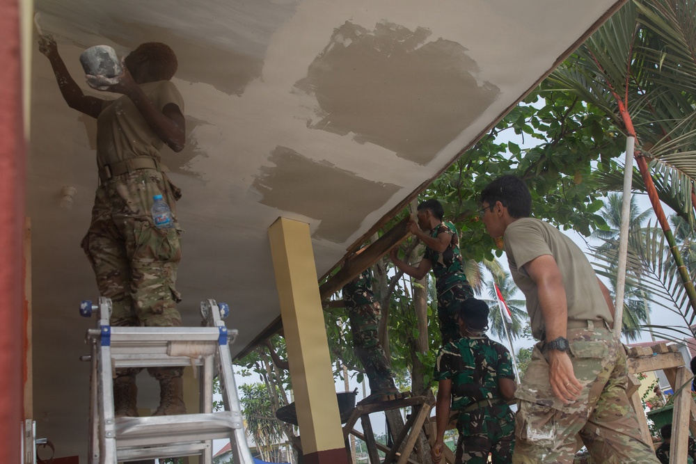 Military personnel in mixed uniforms are engaged in construction or renovation work on a building, plastering or painting a ceiling and repositioning structural panels. The group includes individuals in US-style multicam/OCP camouflage and others in Indonesian TNI (Tentara Nasional Indonesia) woodland camouflage uniforms, suggesting a joint military humanitarian or civic action project. The work is taking place in a tropical environment, likely in Indonesia.
