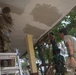 Military personnel in mixed uniforms are engaged in construction or renovation work on a building, plastering or painting a ceiling and repositioning structural panels. The group includes individuals in US-style multicam/OCP camouflage and others in Indonesian TNI (Tentara Nasional Indonesia) woodland camouflage uniforms, suggesting a joint military humanitarian or civic action project. The work is taking place in a tropical environment, likely in Indonesia.