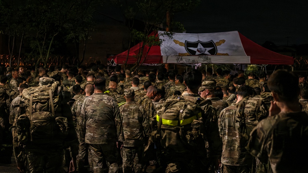 A large formation of military personnel in camouflage uniforms assembled at night in front of a red and white canopy tent bearing a military unit insignia. The gathering appears to be a formal military formation or briefing event, likely associated with the U.S. Army's 2nd Infantry Division based on the visible unit patch on the tent. The scene is dimly lit, suggesting an early morning or nighttime assembly.