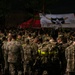 A large formation of military personnel in camouflage uniforms assembled at night in front of a red and white canopy tent bearing a military unit insignia. The gathering appears to be a formal military formation or briefing event, likely associated with the U.S. Army's 2nd Infantry Division based on the visible unit patch on the tent. The scene is dimly lit, suggesting an early morning or nighttime assembly.