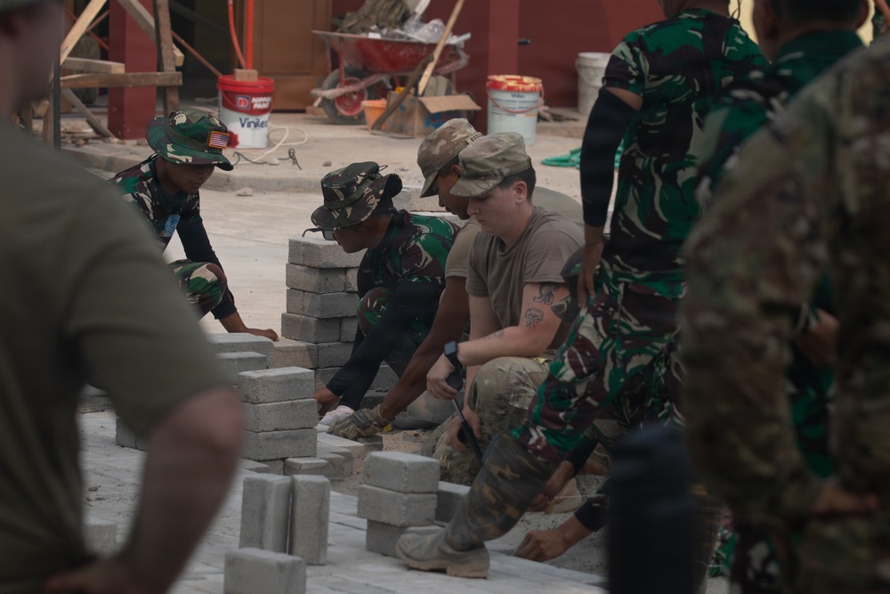 Military personnel from at least two different armed forces are collaborating on a construction project, laying concrete blocks or pavers on a flat surface. One soldier appears to wear a US Army uniform with a tan/multicam cap, while others wear a distinctive green camouflage pattern consistent with Indonesian Armed Forces (TNI) uniforms. The scene takes place at an active construction site with building materials and equipment visible in the background.