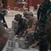 Military personnel from at least two different armed forces are collaborating on a construction project, laying concrete blocks or pavers on a flat surface. One soldier appears to wear a US Army uniform with a tan/multicam cap, while others wear a distinctive green camouflage pattern consistent with Indonesian Armed Forces (TNI) uniforms. The scene takes place at an active construction site with building materials and equipment visible in the background.
